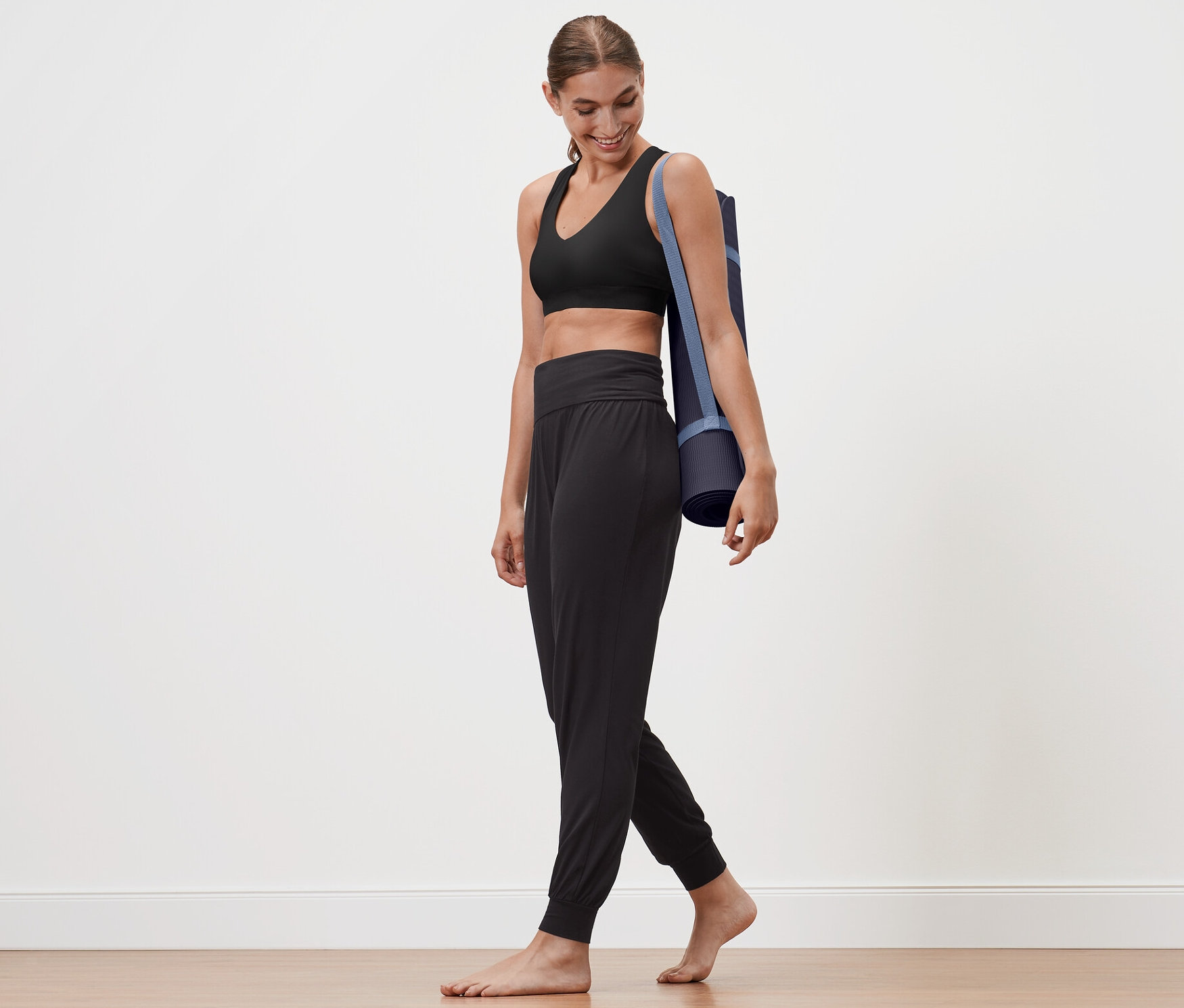 Žena kráčí a nese podložku na jógu přes rameno. Má na sobě černé kalhoty na jógu.