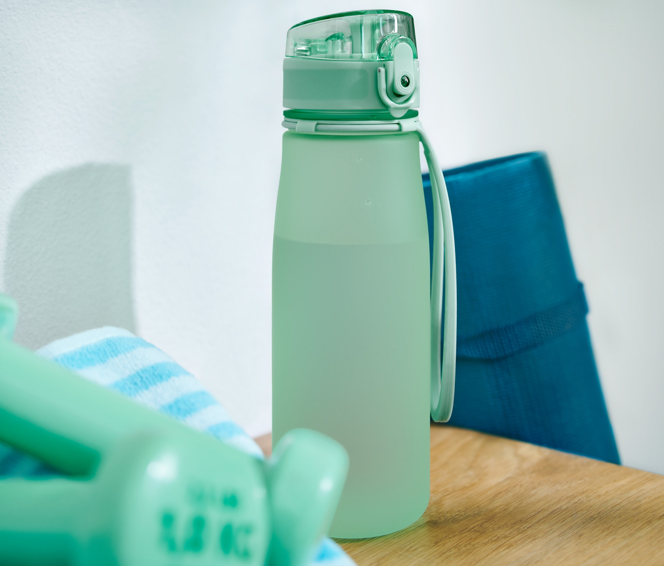 Zelená sportovní láhev na pití stojí na dřevěném stole vedle ručníku, činek a modré podložky na cvičení.