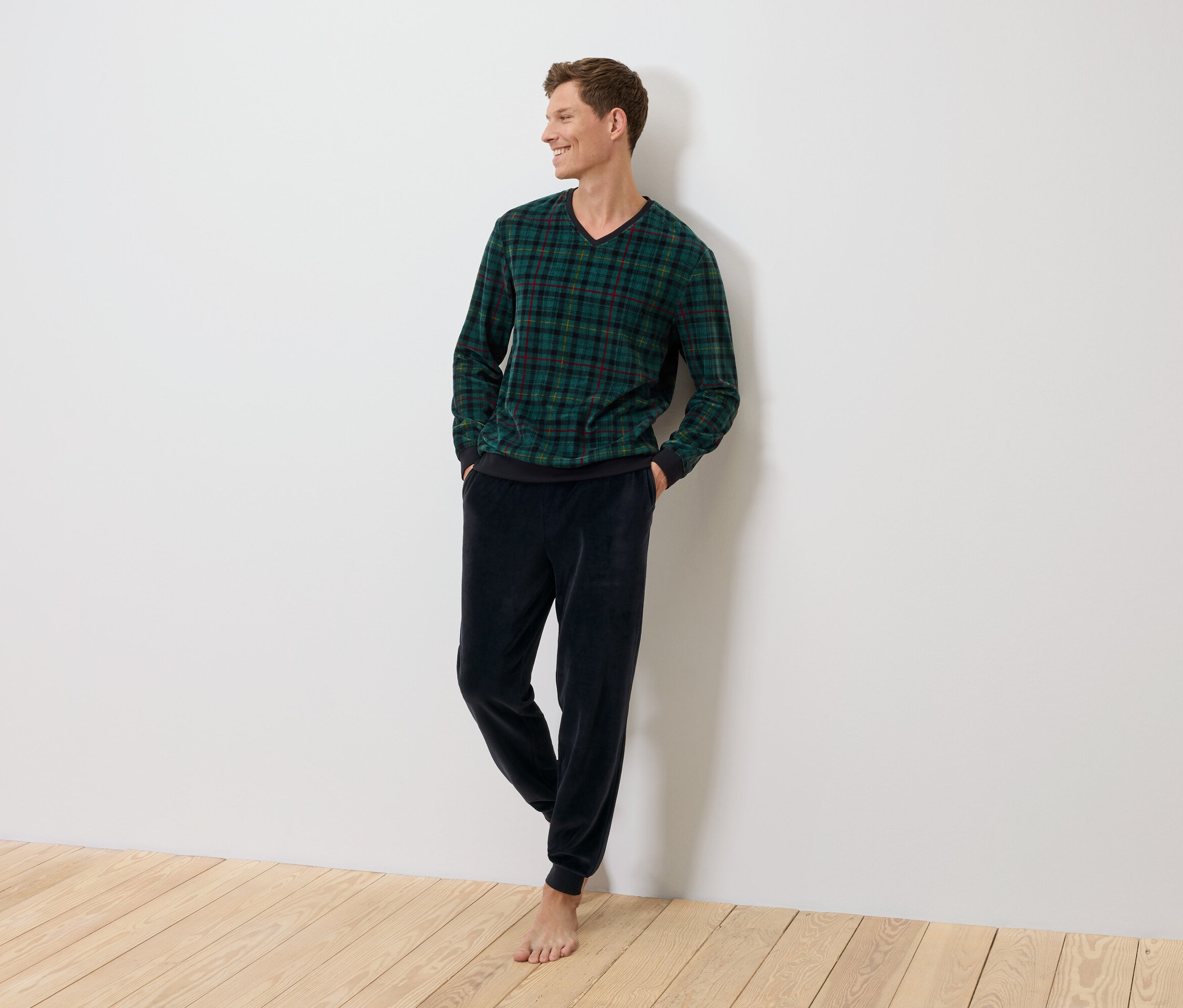 Muž stojí a opírá se o zeď, oblečený do zeleného kostkovaného svetru a tmavě modrých sametových kalhot.