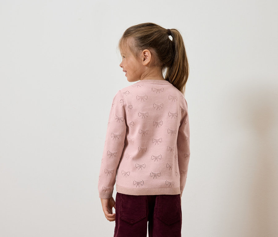 Dívka v růžovém svetru s mašlemi a vínových kalhotách stojí bokem.