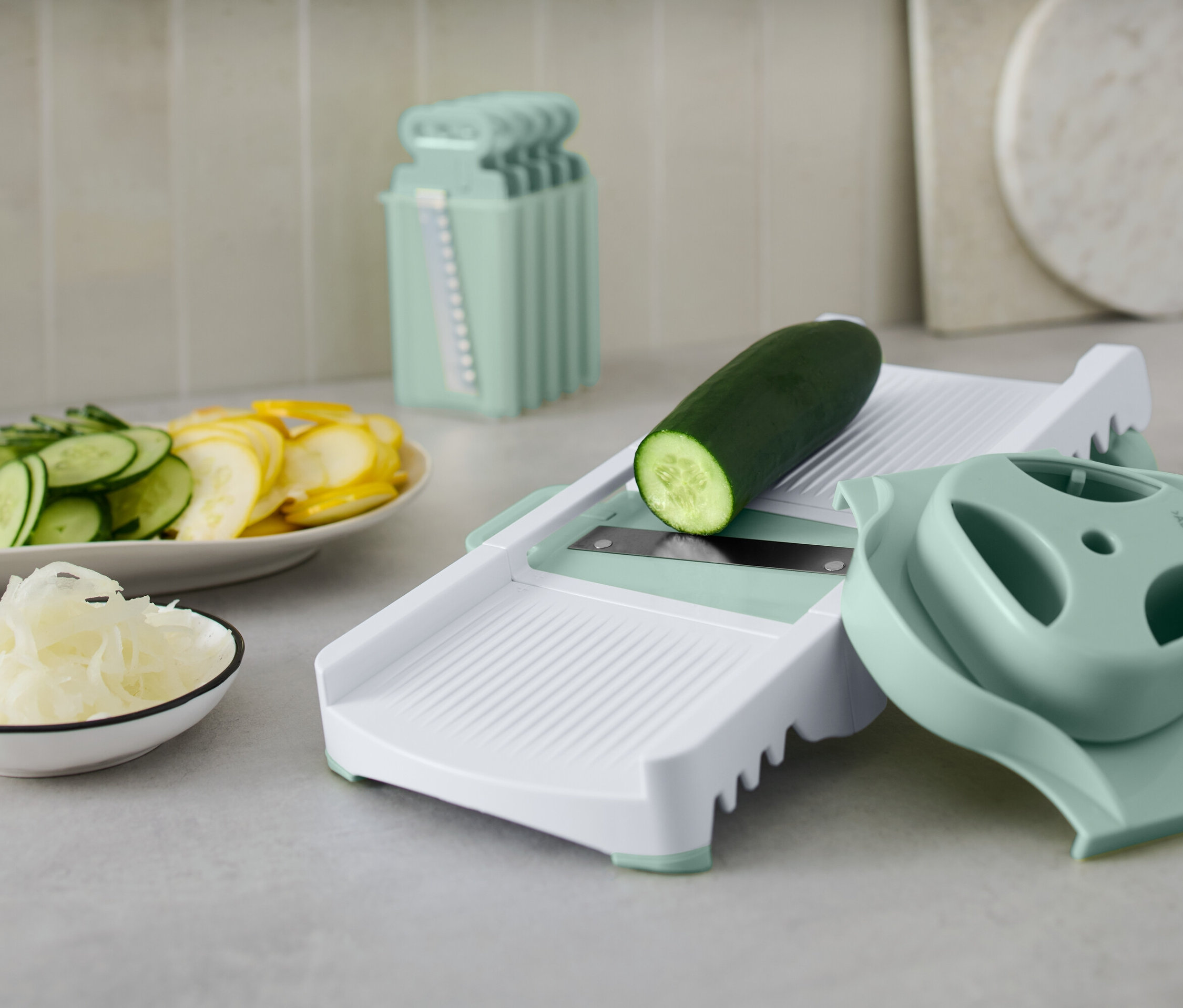 Zelená okurka na bílém kráječi zeleniny s příslušenstvím. Na talíři jsou nakrájené okurky a žlutá tykev.