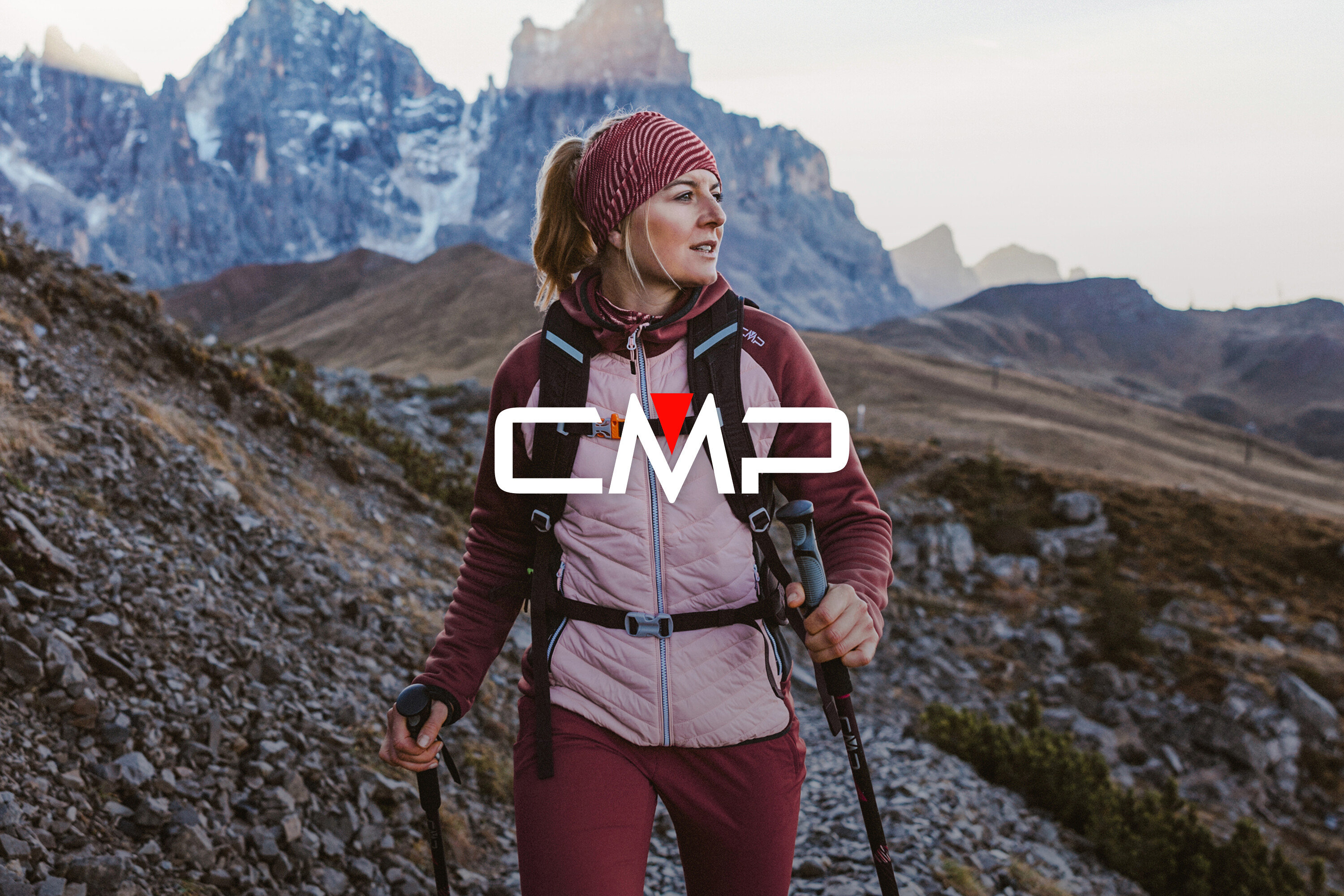 Žena s čelenkou, fleecovou bundou, prošívanou vestou a trekingovými holemi na pozadí hor.