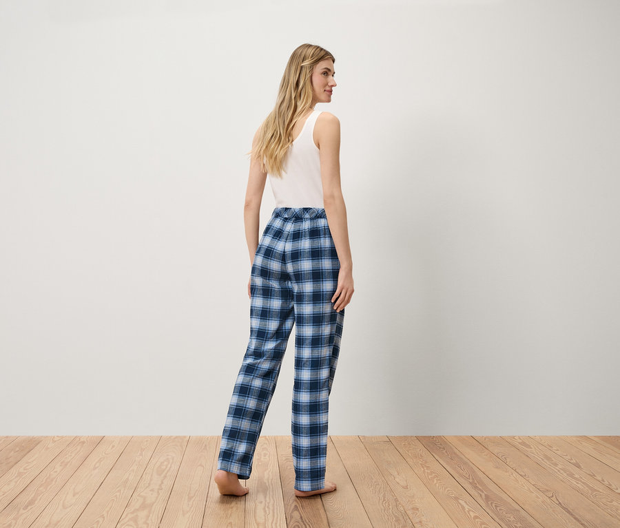 Žena v bílém tílku a modrých kostkovaných kalhotách stojí na dřevěné podlaze.