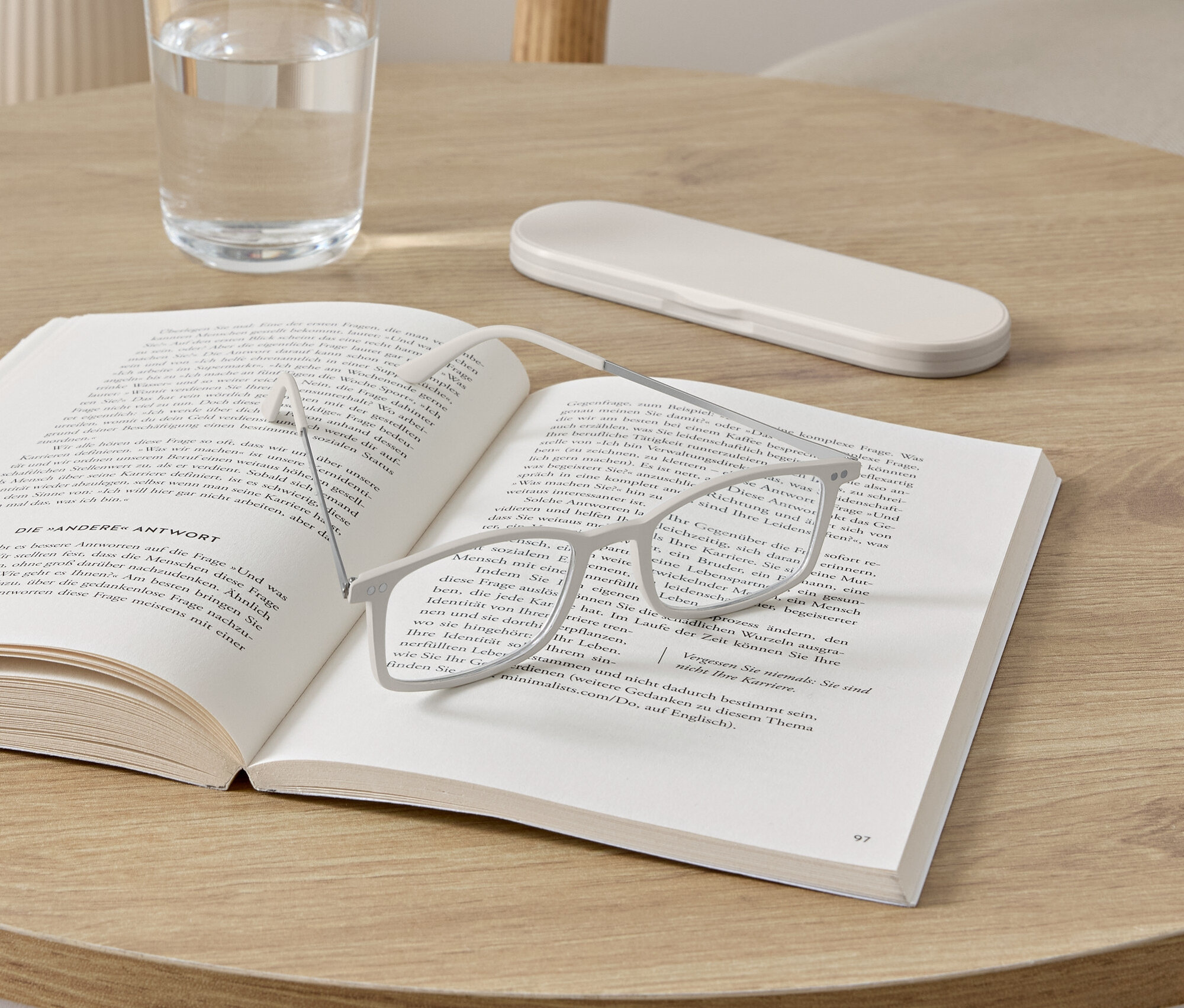 Brýle leží na otevřené knize na kulatém stolku. Vedle knihy stojí sklenice s vodou a pouzdro na brýle.