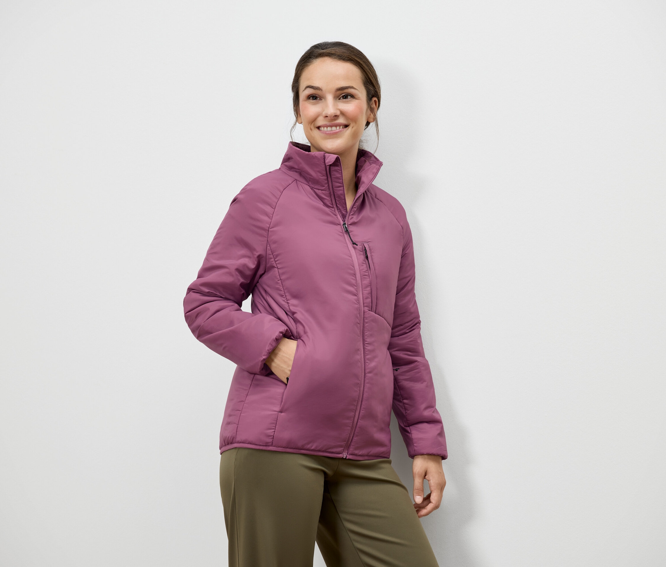 Žena s hnědými vlasy a úsměvem pózuje v růžové bundě a zelených kalhotách před bílou zdí.