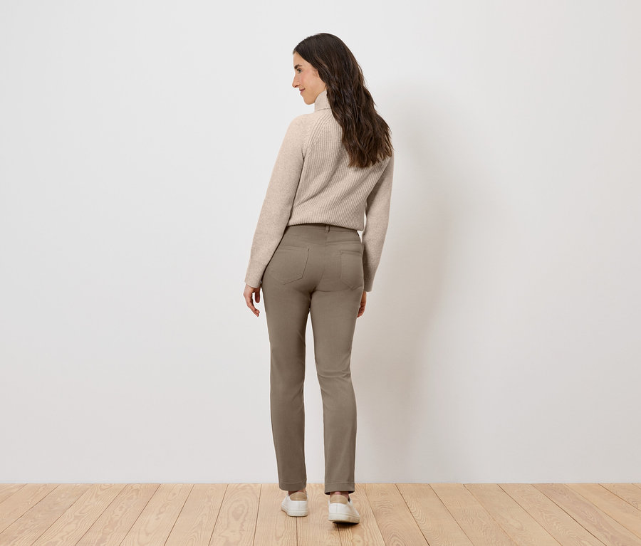 Žena v béžovém svetru a hnědých kalhotách stojící zády k divákovi.