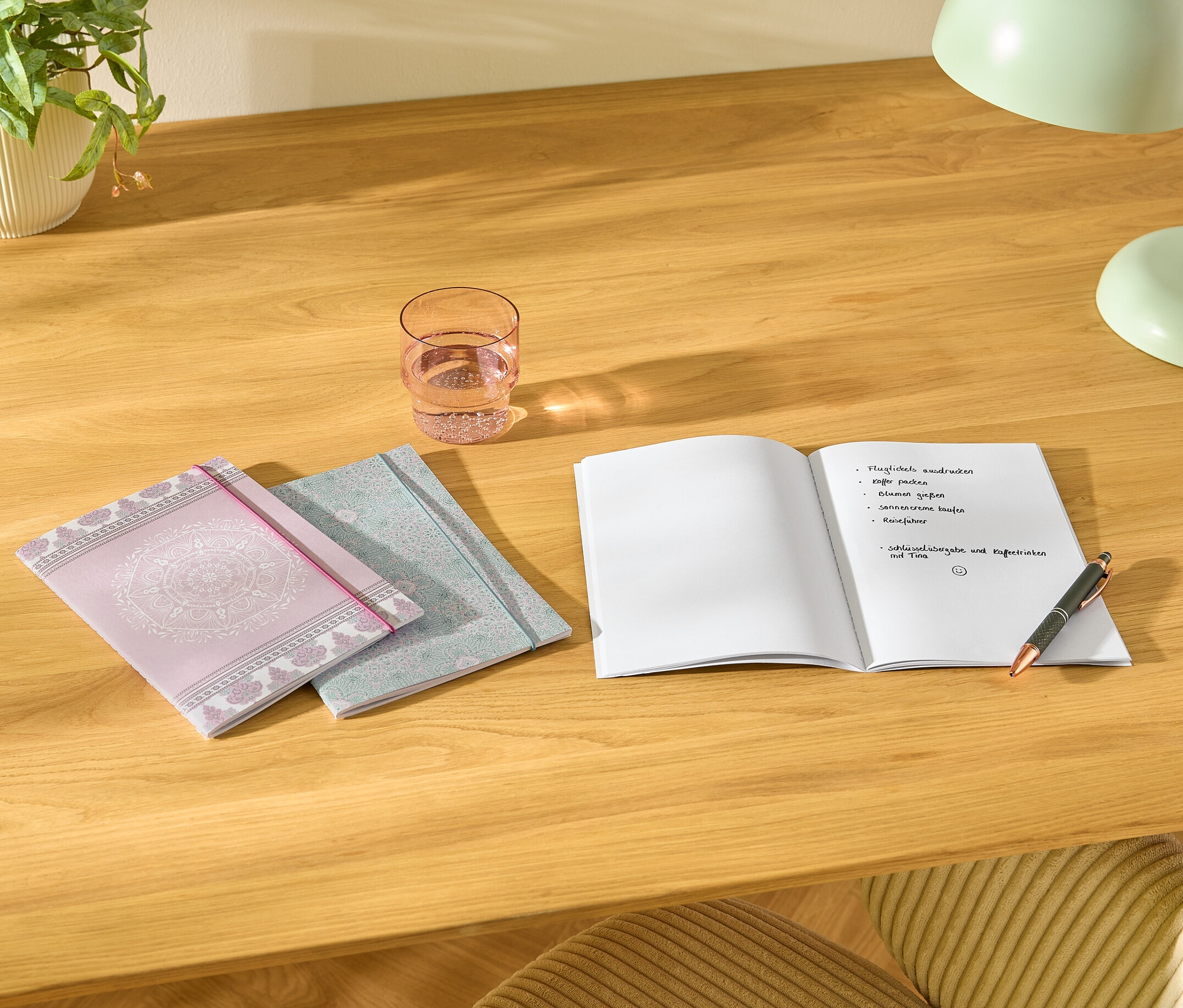 Na stole leží tři poznámkové bloky, sklenice vody, otevřený zápisník s poznámkami a pero.