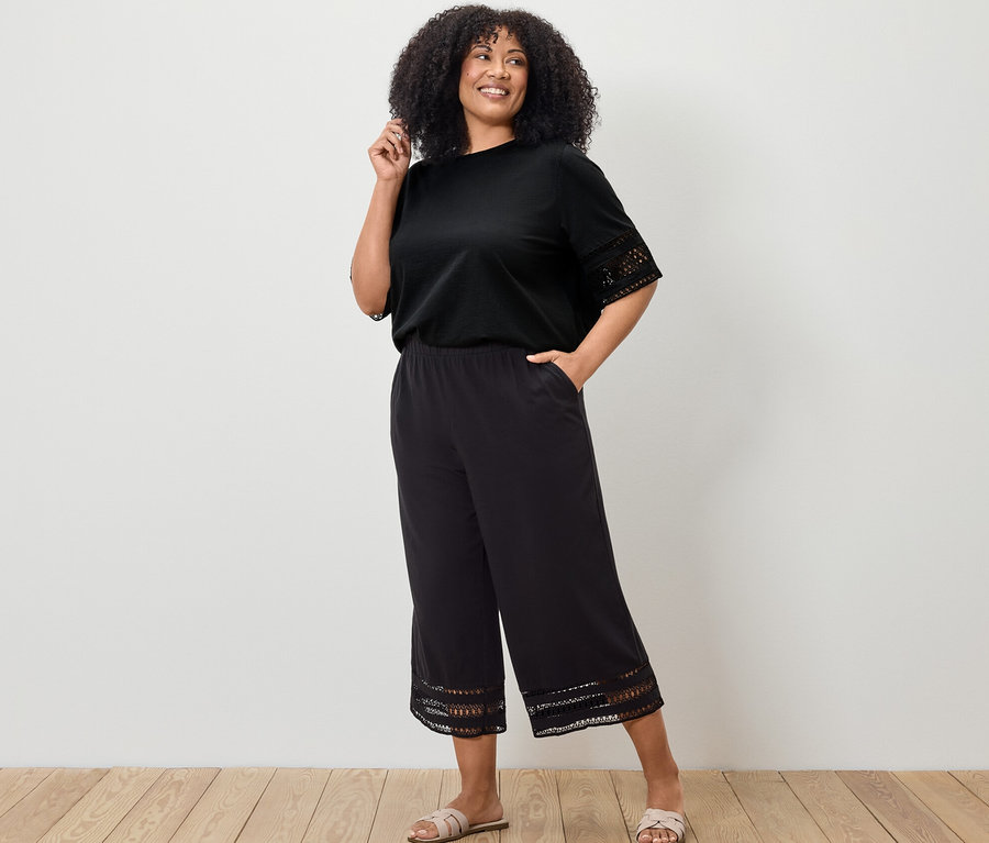 Žena pózuje v černém tričku s výšivkou a černých culotte kalhotách s výšivkou.