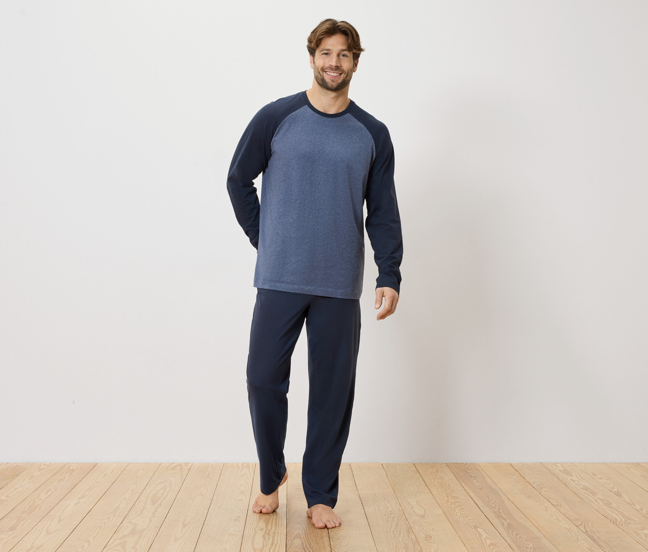 Muž stojí bosý na dřevěné podlaze a má na sobě tmavě modré pyžamo.