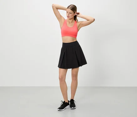 Žena se zvednutýma rukama má na sobě korálový sportovní top, černou sukni a černé tenisky.