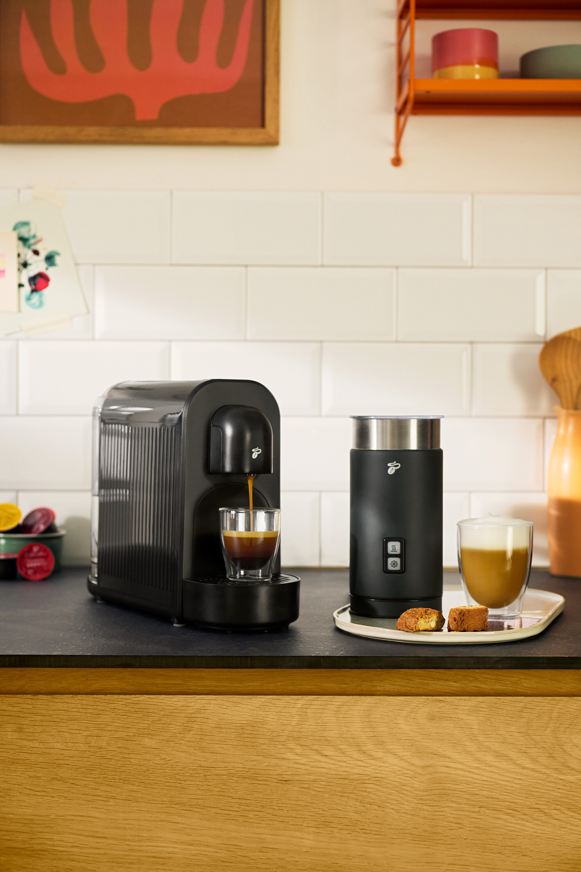 Káva teče z černého kávovaru do sklenice. Vedle stojí černý elektrický napěňovač mléka.