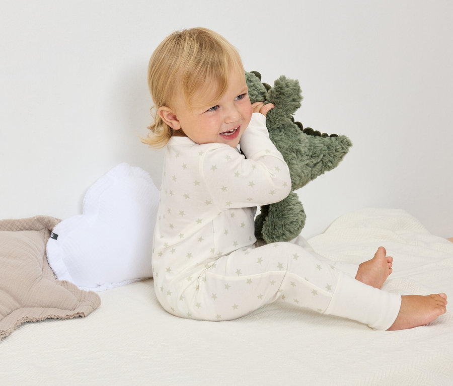 Blonďaté dítě v bílém hvězdičkovaném pyžamu sedí na bílé dece a objímá zeleného plyšového dinosaura.