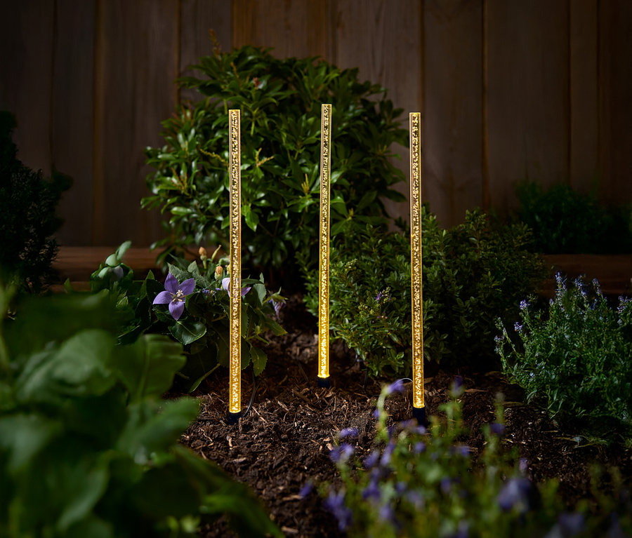 Tři zlaté solární zahradní lampy stojí na záhonu s keři a květinami.