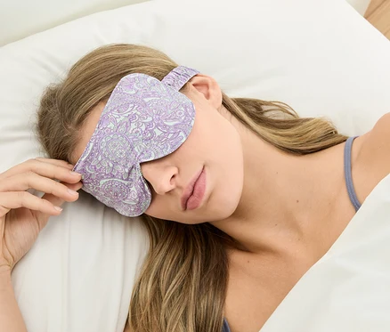 Žena leží na bílém polštáři s fialovou maskou na spaní se vzorem.
