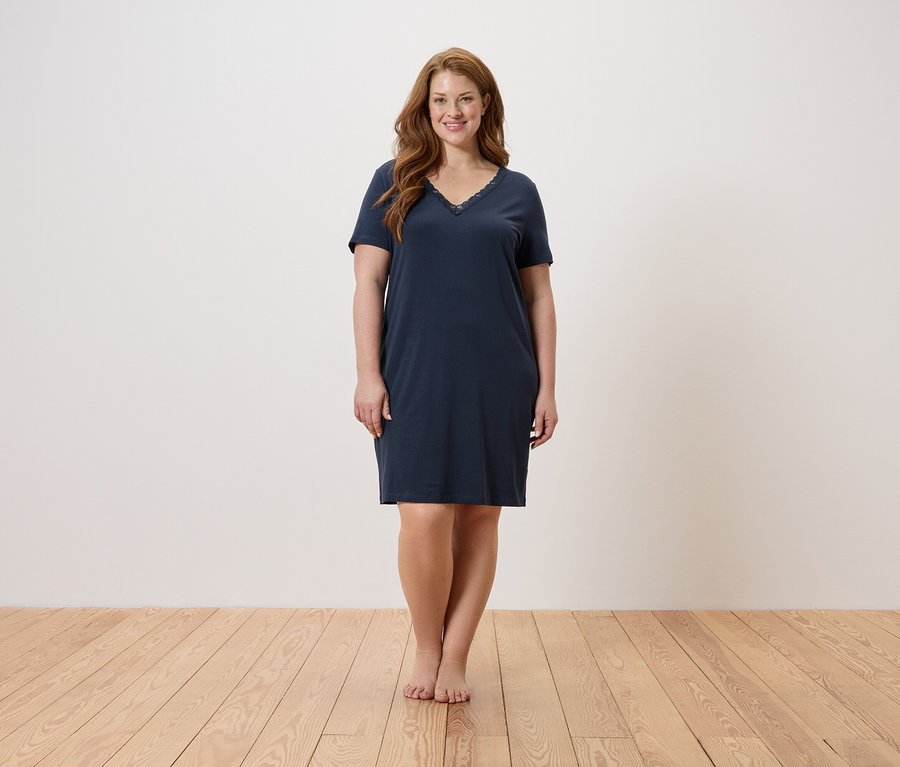 Žena stojí bosá na dřevěné podlaze a má na sobě tmavě modrou noční košili.