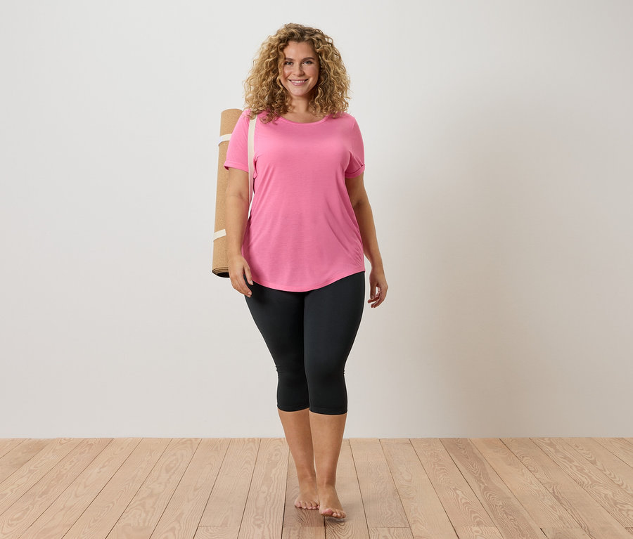 Žena s kudrnatými vlasy má na sobě růžové tričko a černé legíny, s podložkou na jógu přes rameno, bosá na dřevěné podlaze.