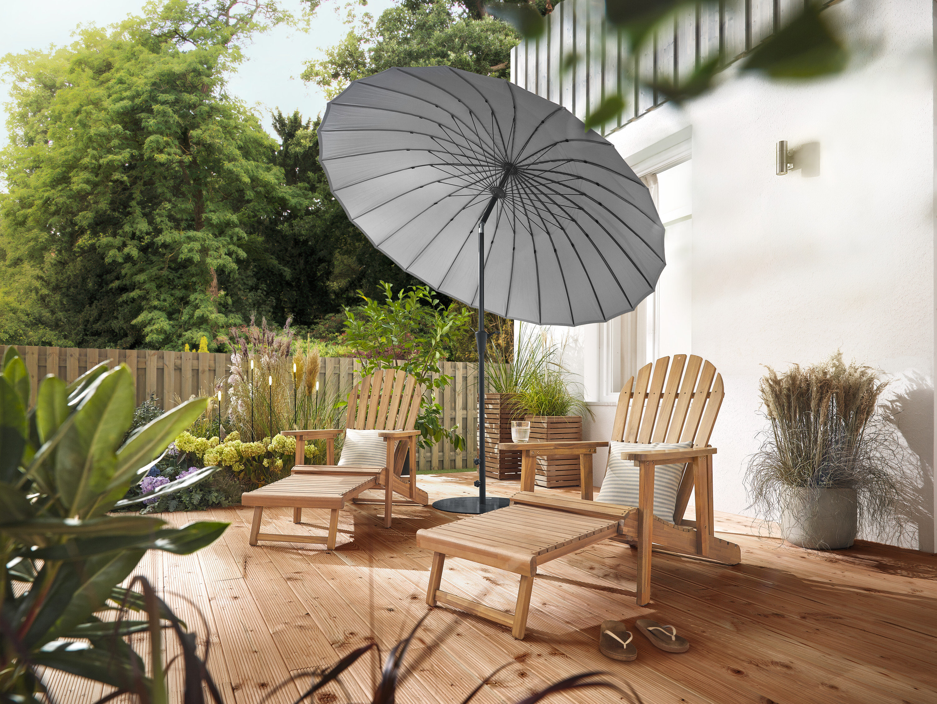 Na terase stojí dvě přírodní zahradní křesla Adirondack s výsuvnou podnožkou, lemované 2 Pflanzsäulen »Tinus« a 5 LED solárními zápichy. Světle šedý slunečník Leco Shanghai poskytuje stín.