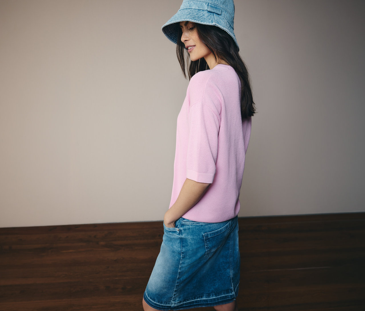 Žena s džínovým kloboukem, růžovým svetrem s krátkým rukávem CECIL a džínovou sukní CECIL. Má ruce v kapsách.