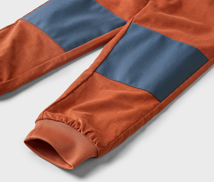 Detail oranžových manšestrových kalhot s modrými záplatami na kolenou a elastickými manžetami na kotnících.