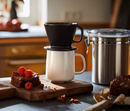 Černý ruční kávový filtr velikosti 101 stojí na bílém hrnku vedle brownies s malinami na dřevěném prkénku.