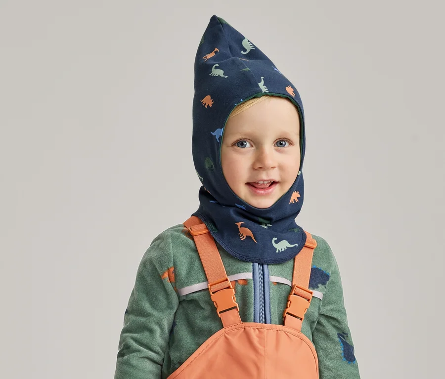 Usmívající se chlapec v modré kukle s dinosaury, zelené fleecové mikině a oranžových lacláčích na šedém pozadí.