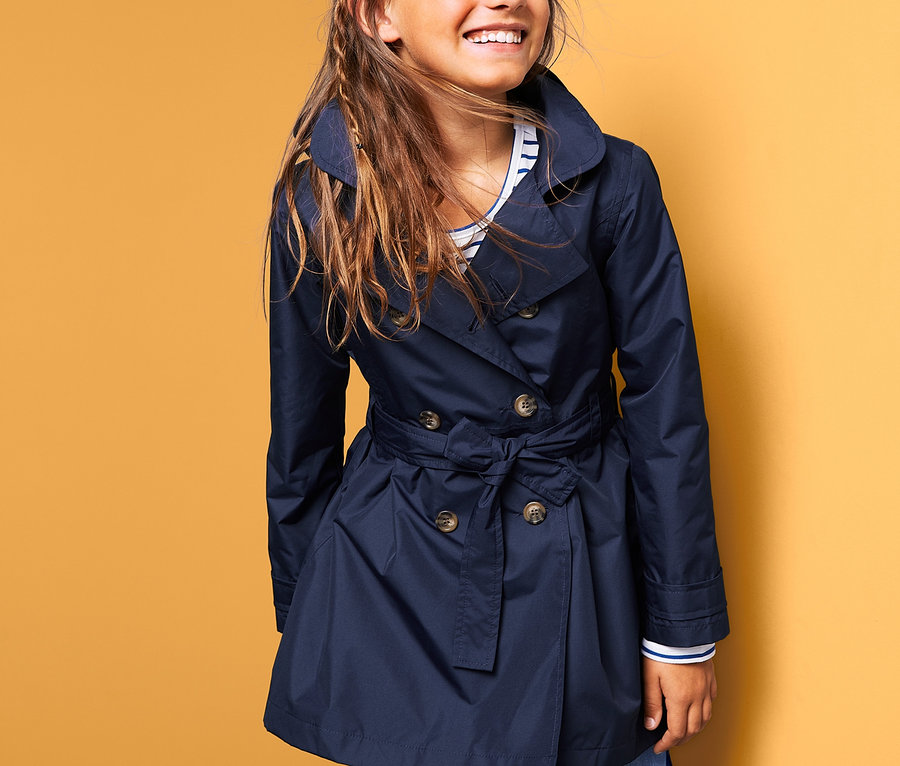 Usmívající se dívka má na sobě tmavě modrý trenčkot do každého počasí.