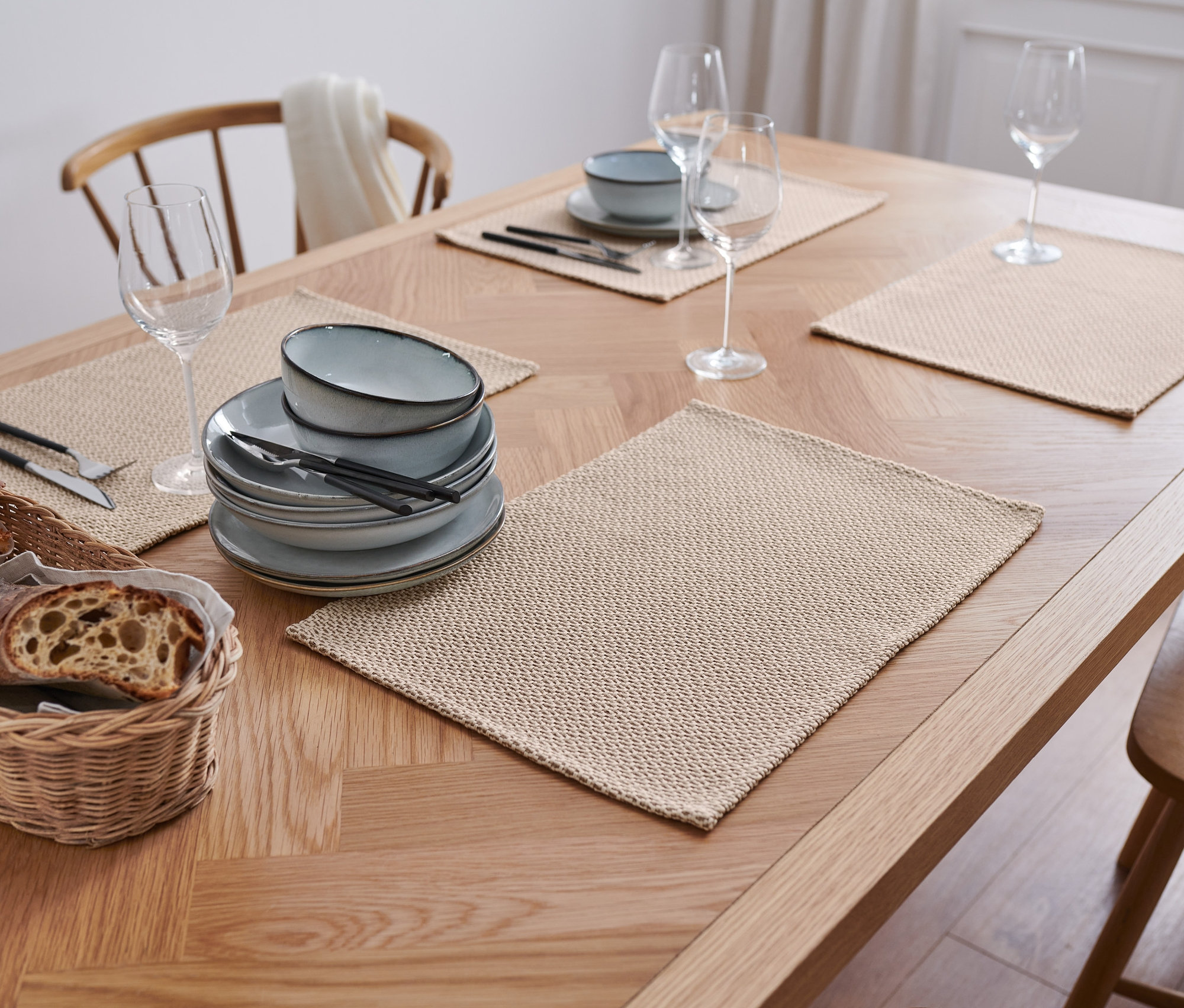 Na dřevěném stole leží 4 béžová prostírání, talíře, příbory, sklenice a košík s chlebem.
