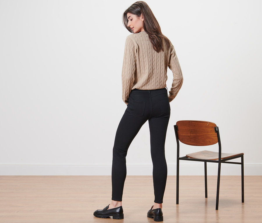 Žena stojí zády, má na sobě béžový svetr a černé džegíny.