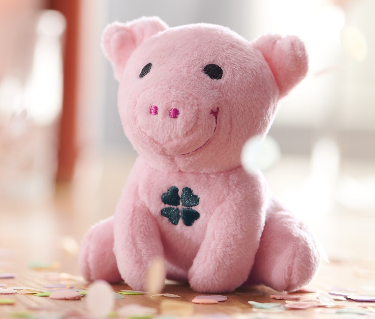 Sedící plyšové chrochtající prasátko pro štěstí s jetelovým listem na břiše, obklopené konfetami.