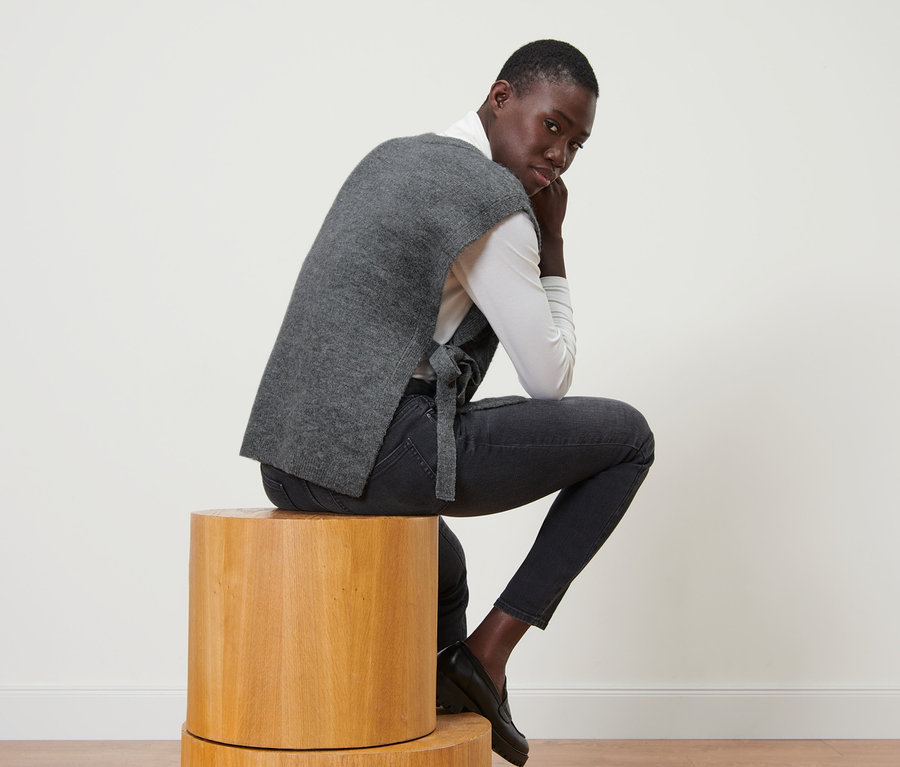 Muž sedí na dřevěné stoličce a má na sobě pletenou vestu se šňůrkami na zavazování.