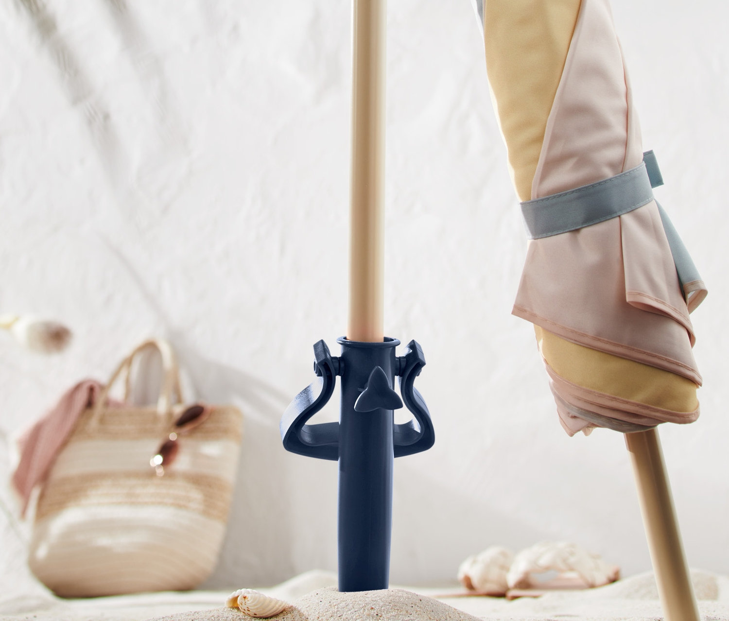 Pastelově pruhovaný slunečník je upevněn v modrém šroubovacím držáku na slunečník na písku.