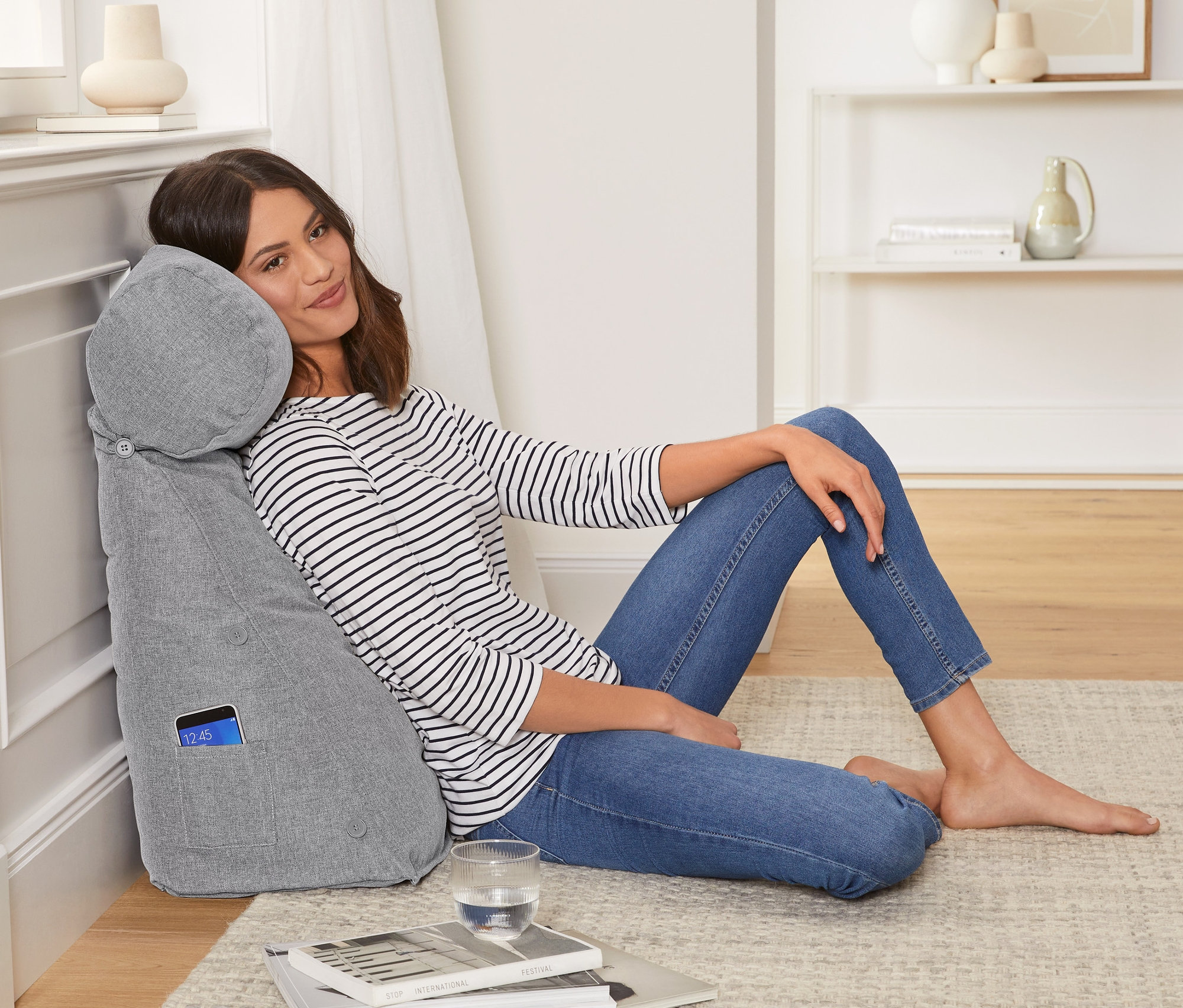 Žena sedí opřená o šedý polštář za záda se šíjovým polštářkem.