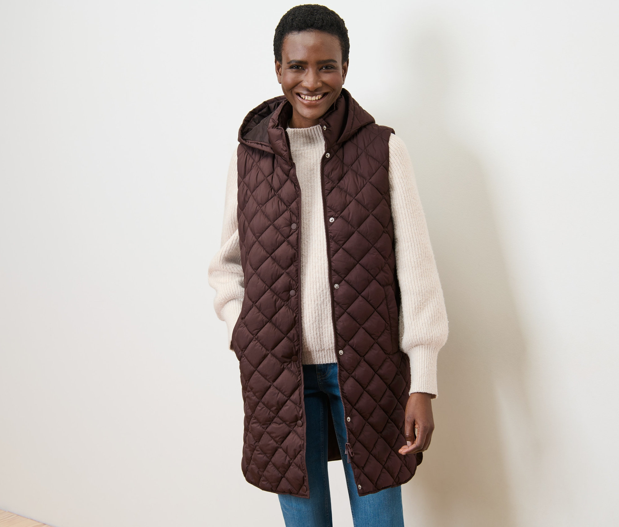 Žena usměvavě pózuje v dlouhé prošívané vestě s kapucí.