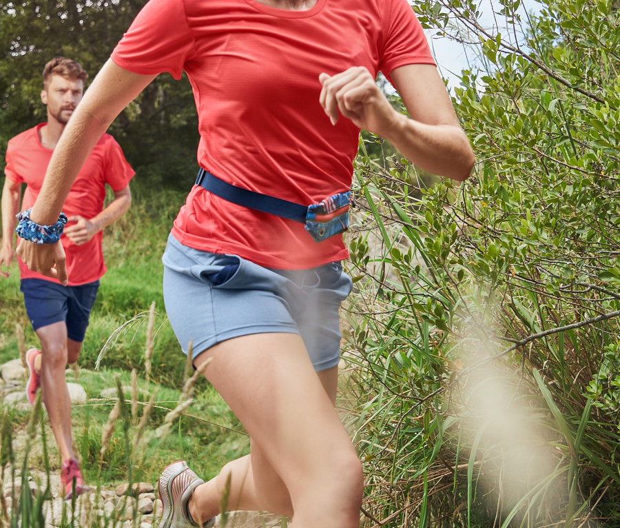 Žena v korálově červeném funkčním tričku a 3/4 sportovních legínách tmavě modré barvy běží po kamenité cestě. V pozadí běží muž v modrých funkčních šortkách a korálově červeném sportovním tričku.