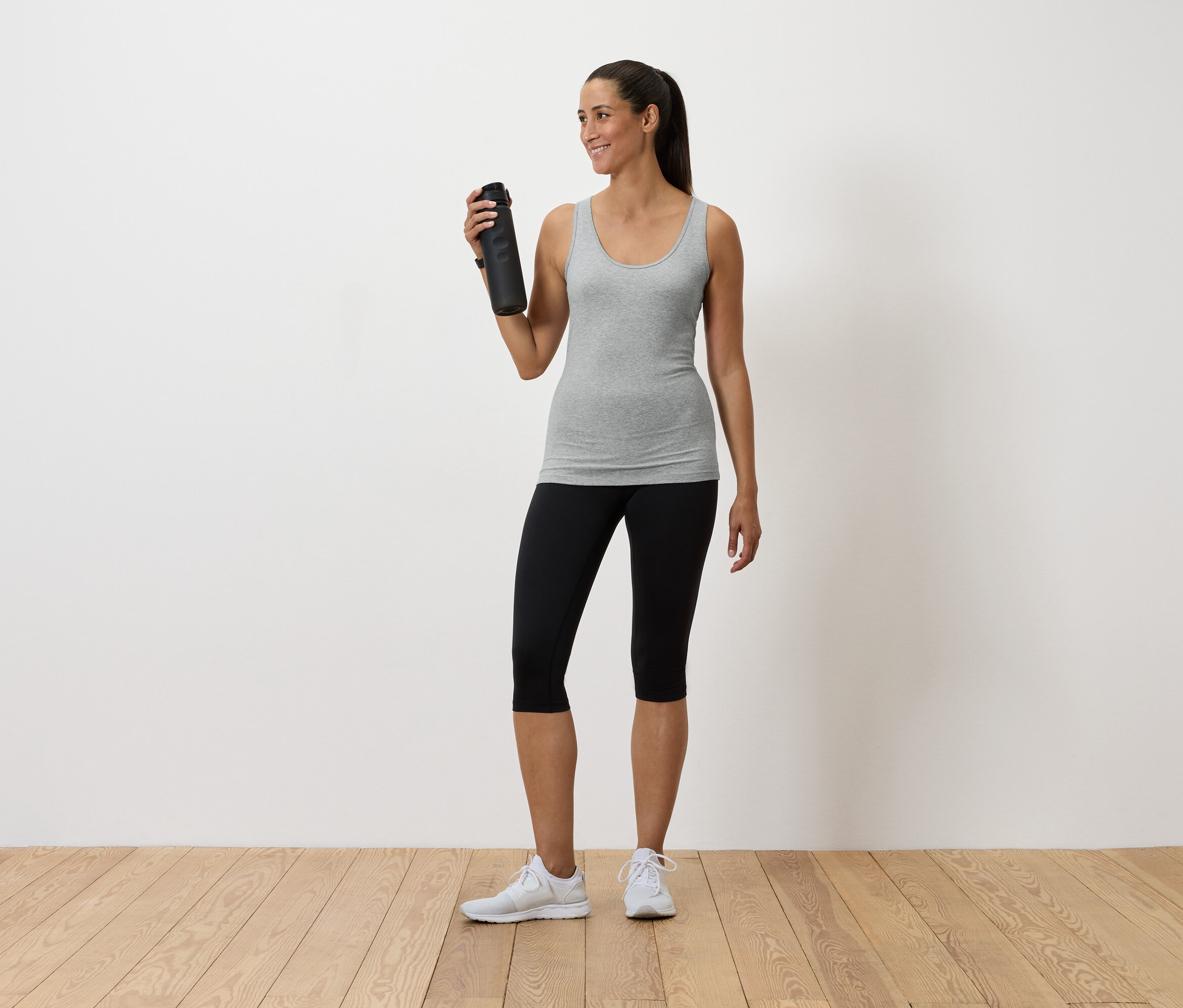 Žena drží v ruce láhev s vodou, má na sobě šedé tílko, černé legíny a bílé tenisky.