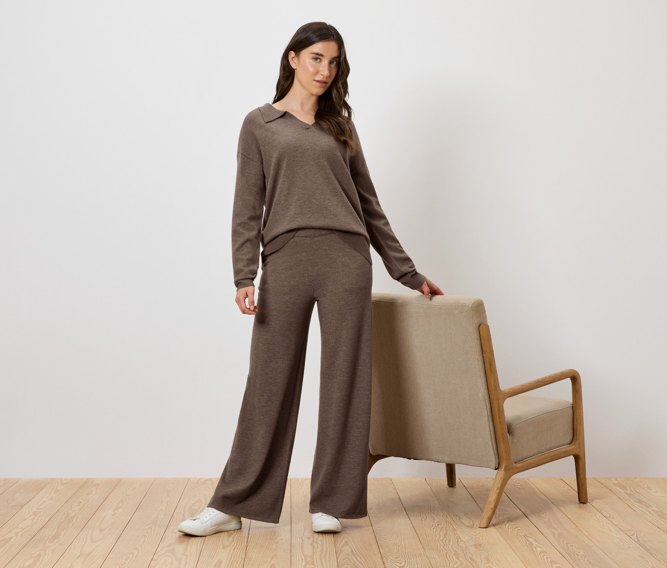 Žena v hnědé pletené soupravě pózuje vedle křesla.