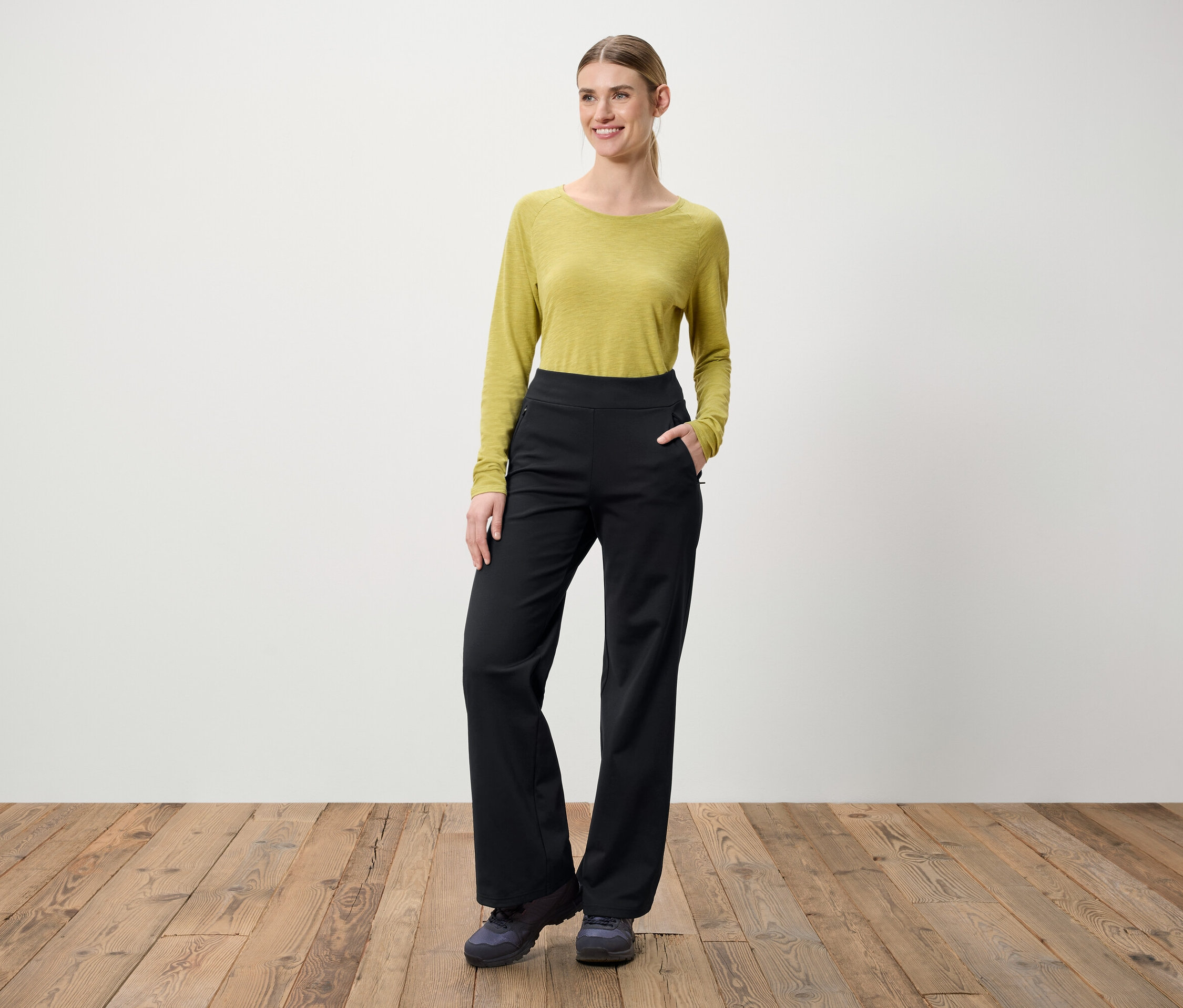 Žena stojí na dřevěné podlaze. Má na sobě zelenou blůzu s dlouhým rukávem a černé kalhoty.