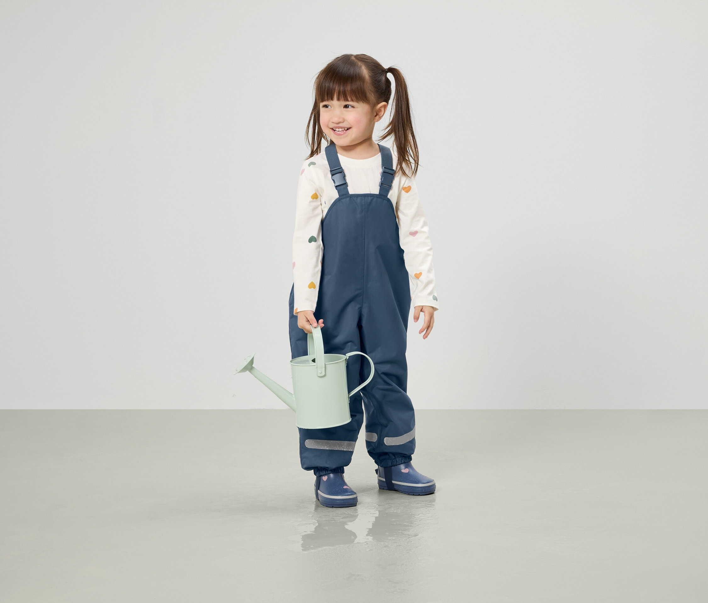 Usmívající se dívka s culíky drží světle zelenou konev, oblečená v modrých kalhotách a botách do deště.