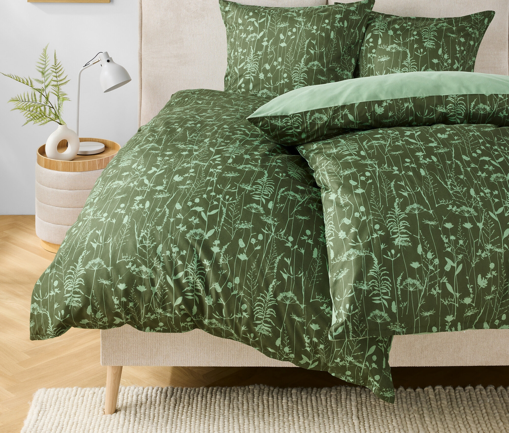 Zelené povlečení s květinovým vzorem na posteli. Na nočním stolku stojí lampa a váza s rostlinou.