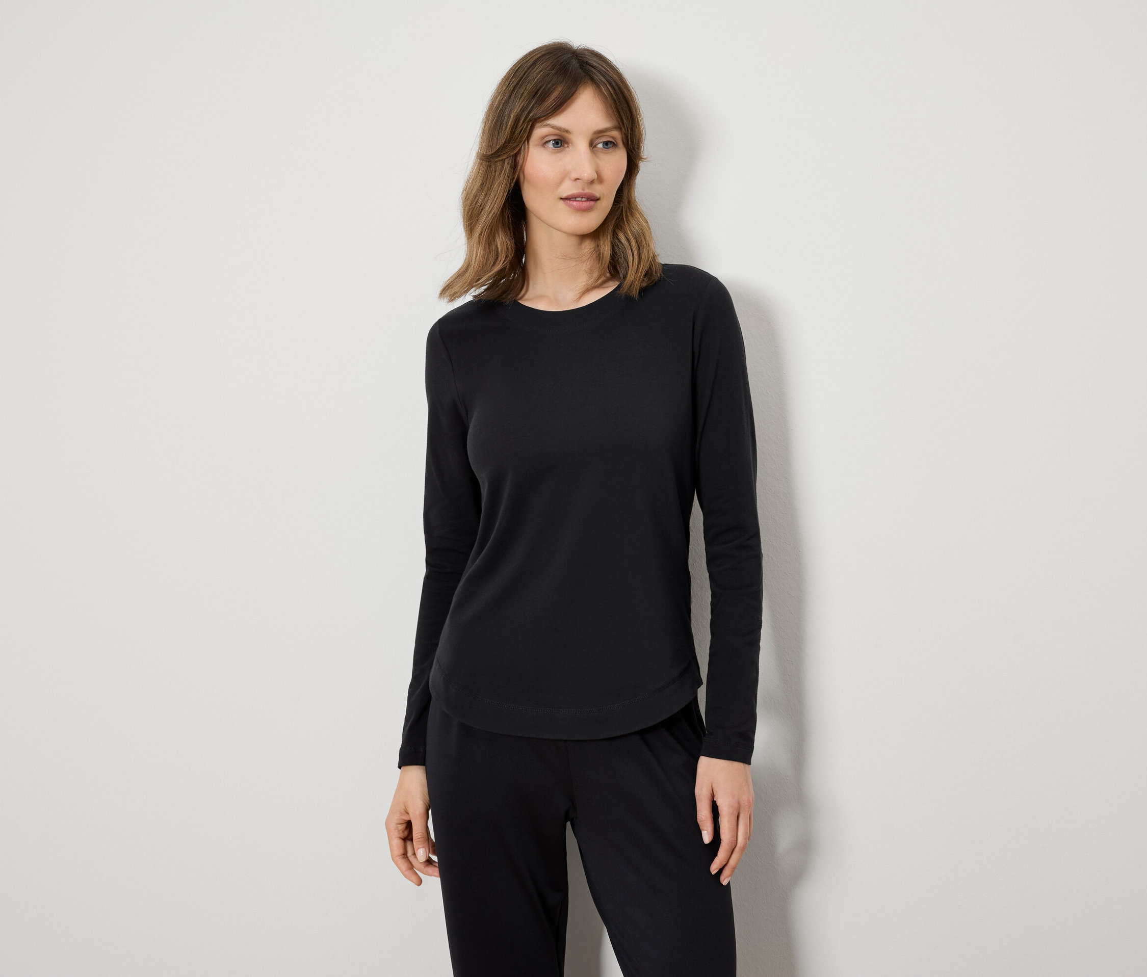 Žena v černém tričku s dlouhým rukávem a černých kalhotách stojí před bílou zdí.