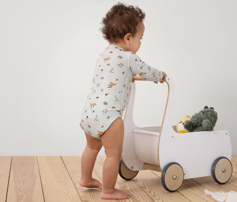 Batole tlačí bílý dřevěný kočárek s hračkami na dřevěné podlaze.