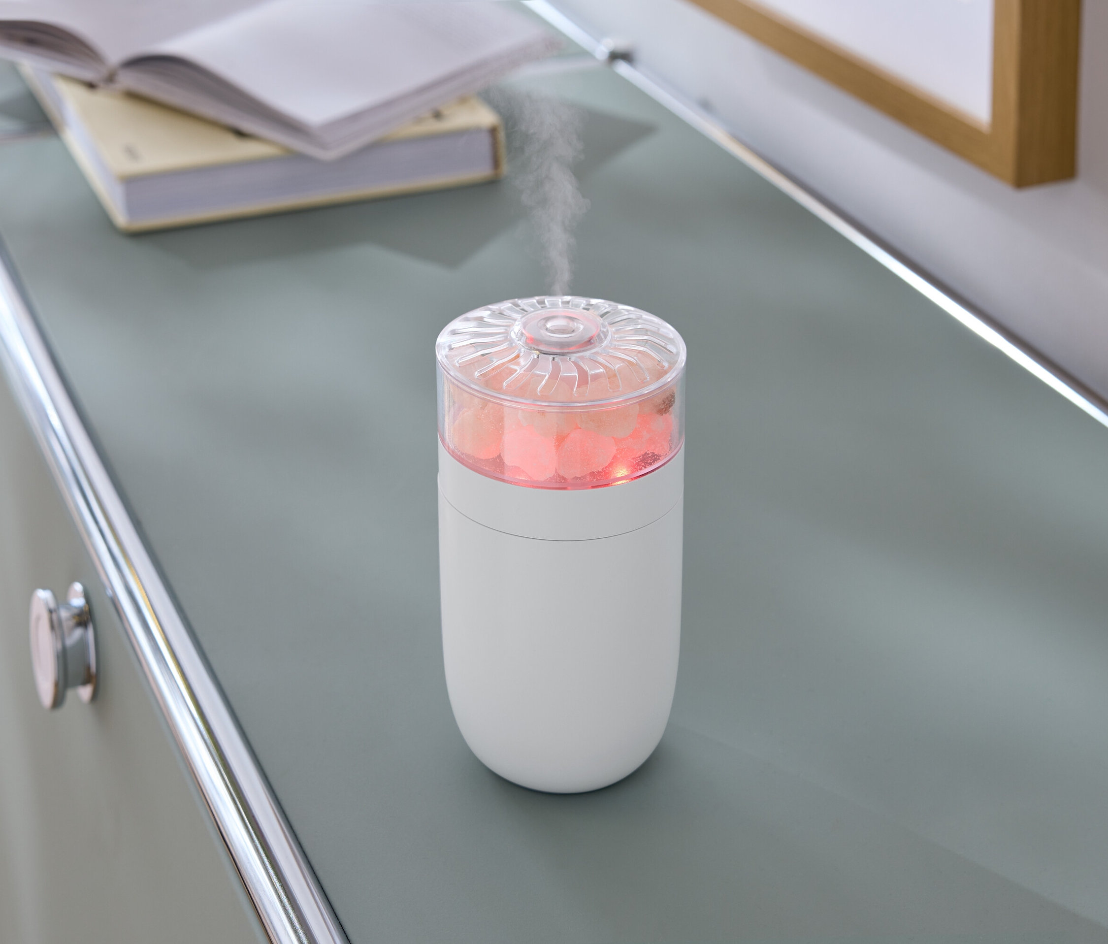 Na šedé komodě stojí bílý zvlhčovač vzduchu s růžovými kameny a párou.