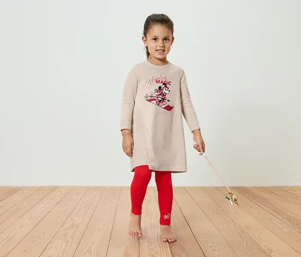 Dívka v béžových šatech s motivem Minnie Mouse a červených legínách stojí na dřevěné podlaze.