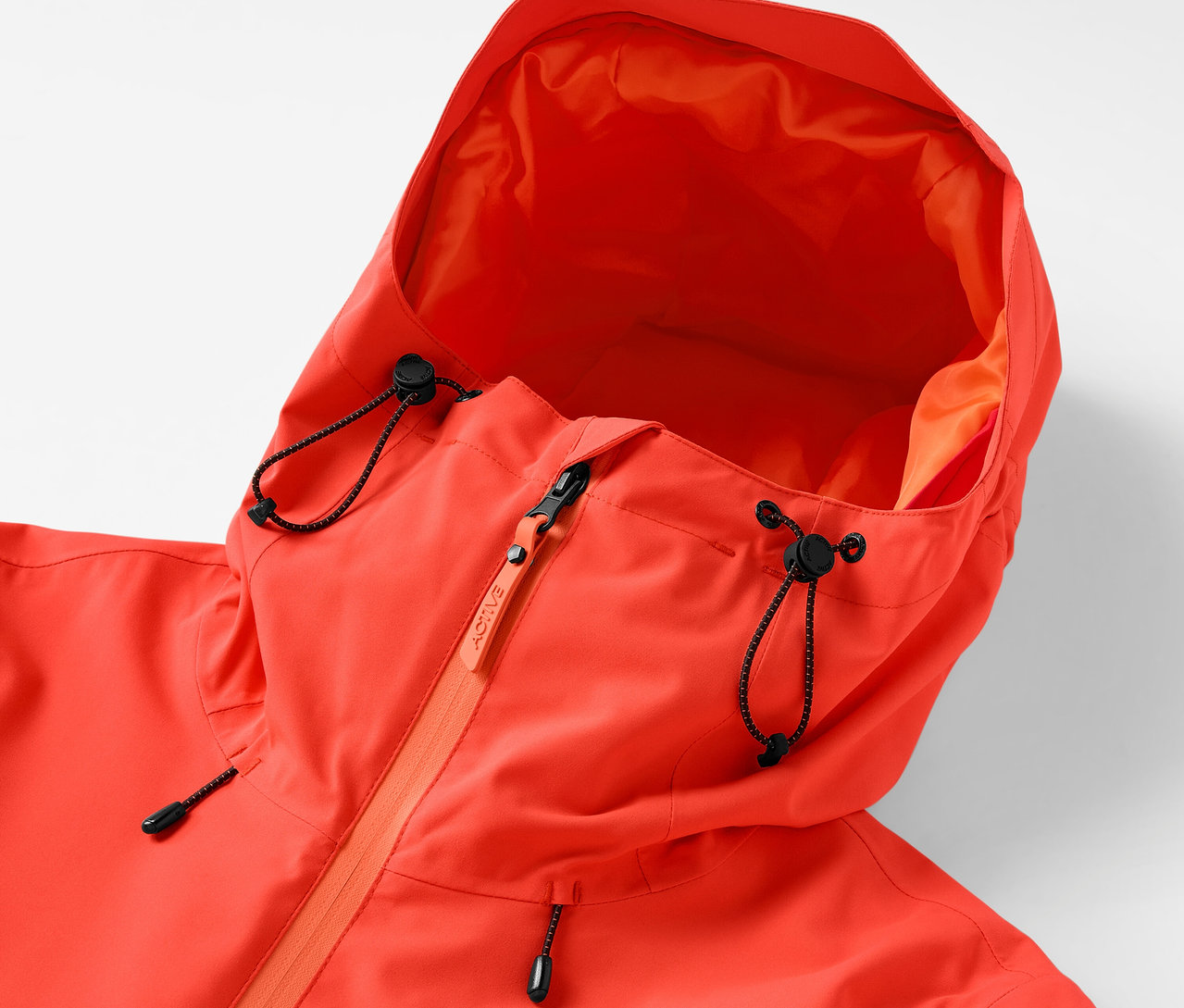 Detail oranžové bundy s kapucí se stahovací šňůrkou a zipem.