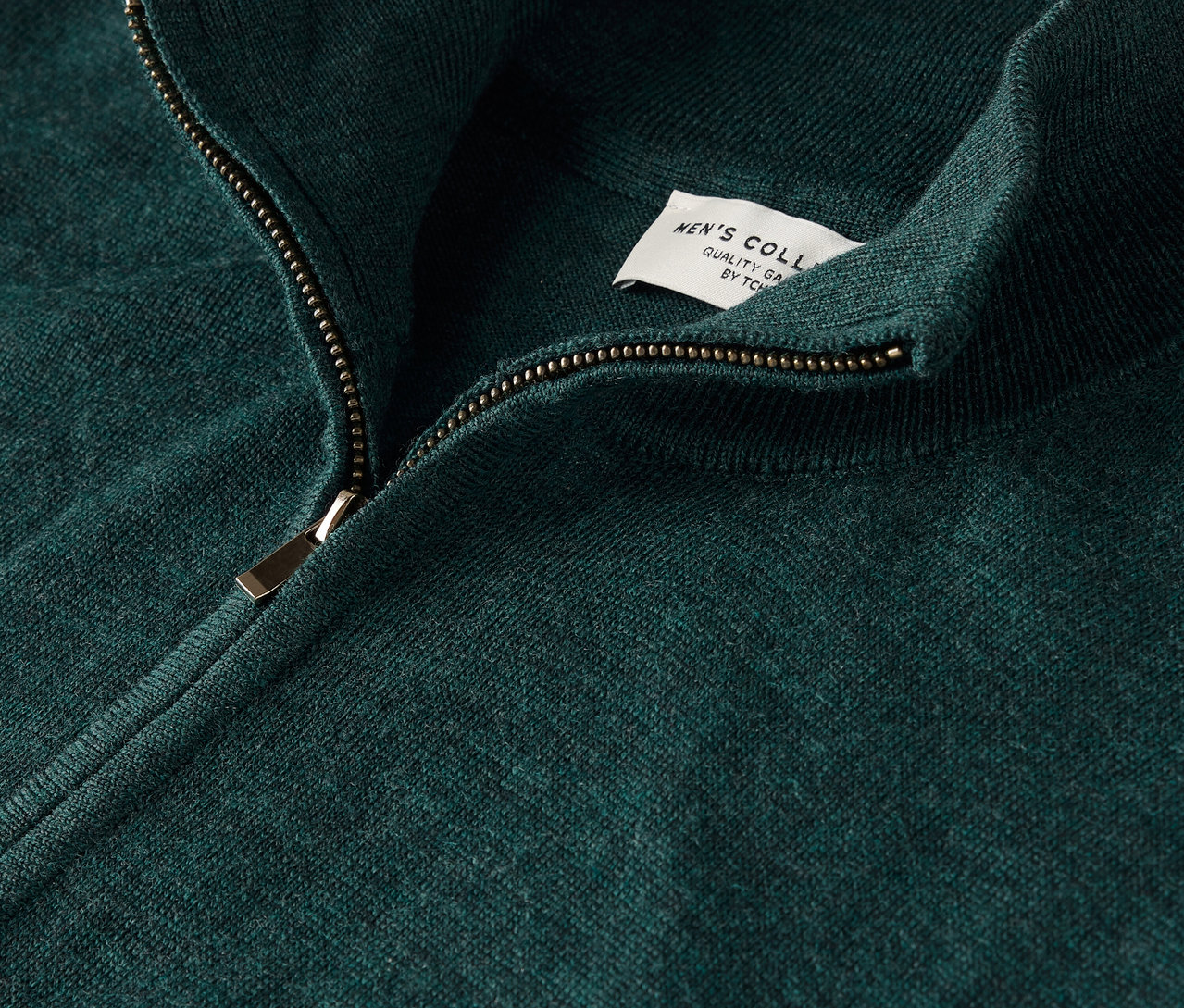 Detail zeleného svetru se zipem a štítkem.