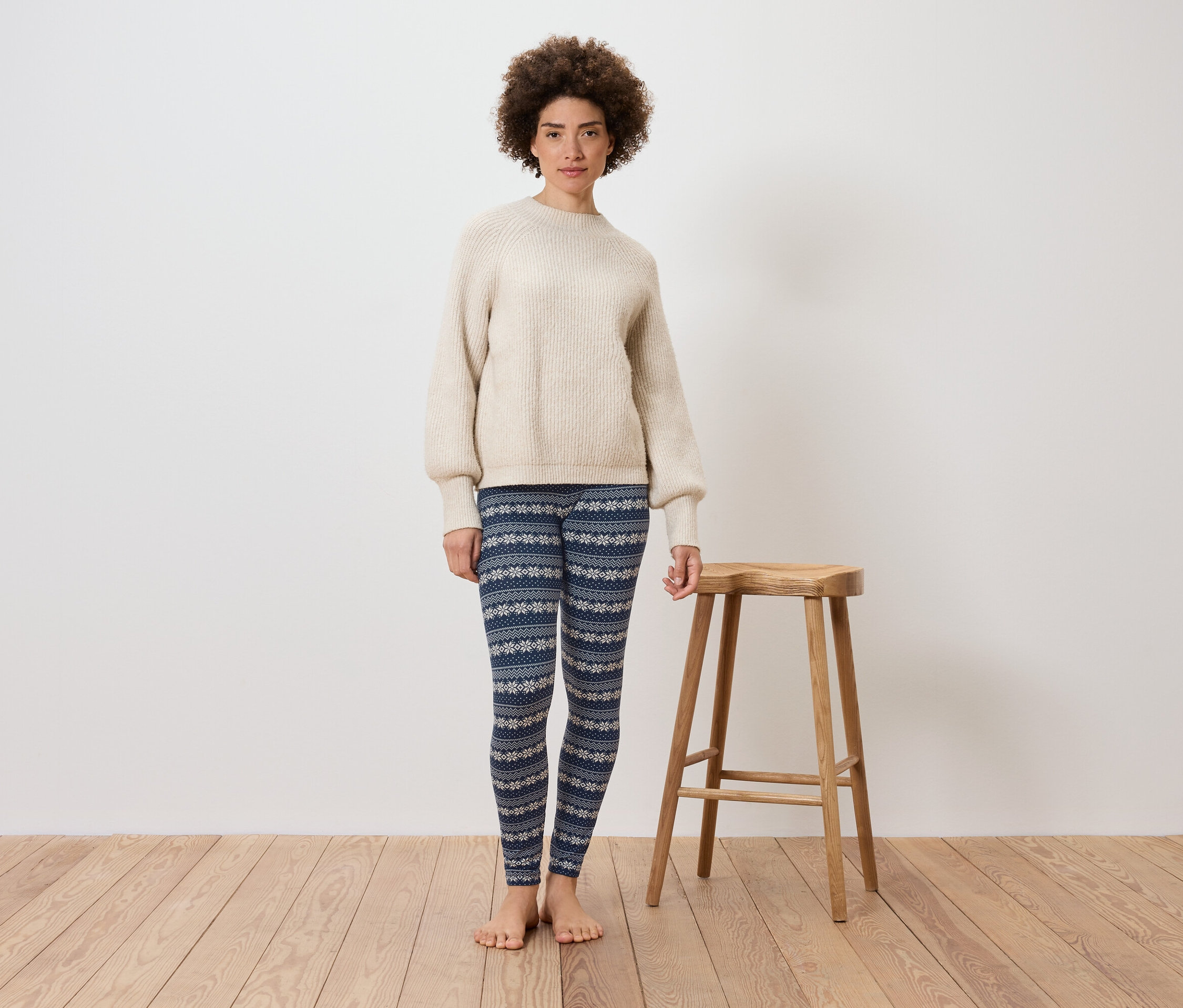 Žena stojí před bílou zdí a vedle dřevěné stoličky. Má na sobě krémový svetr a modré legíny se vzorem.