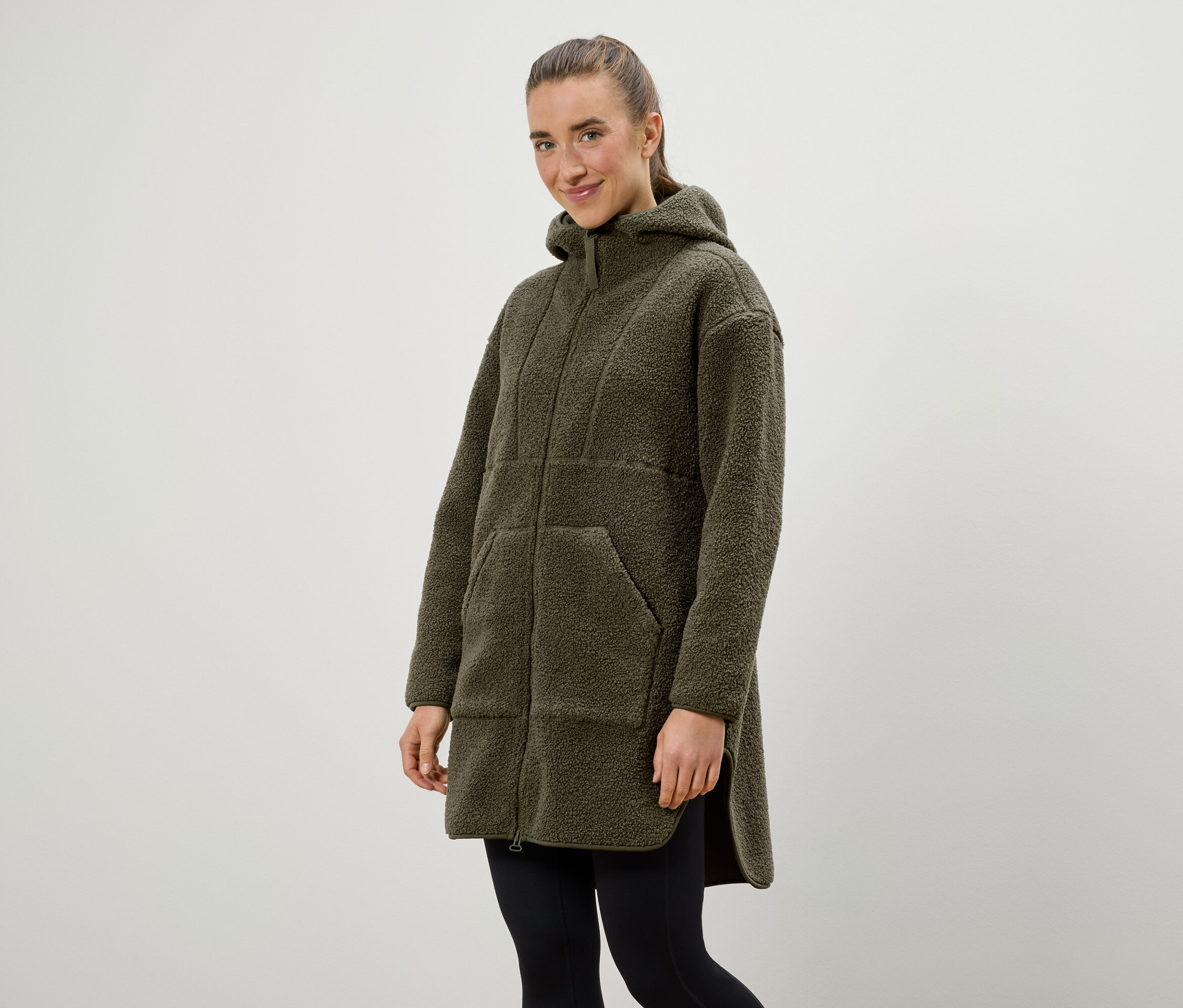 Žena má na sobě olivově zelený kabát s kapucí a černou legíny.