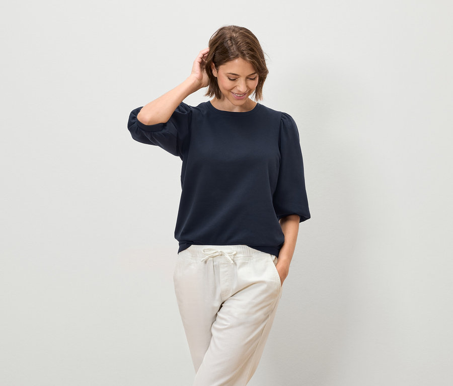 Žena pózuje v tmavě modré haleně s krátkými rukávy a světlých kalhotách.