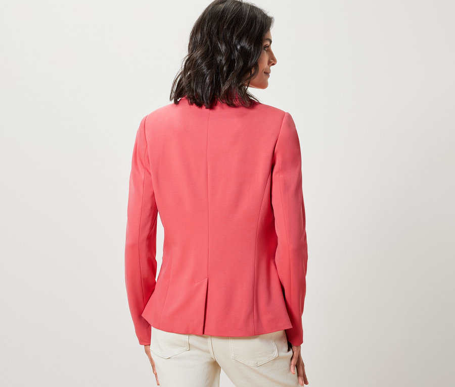 Žena stojí zády, má na sobě korálové sako a světlé kalhoty.