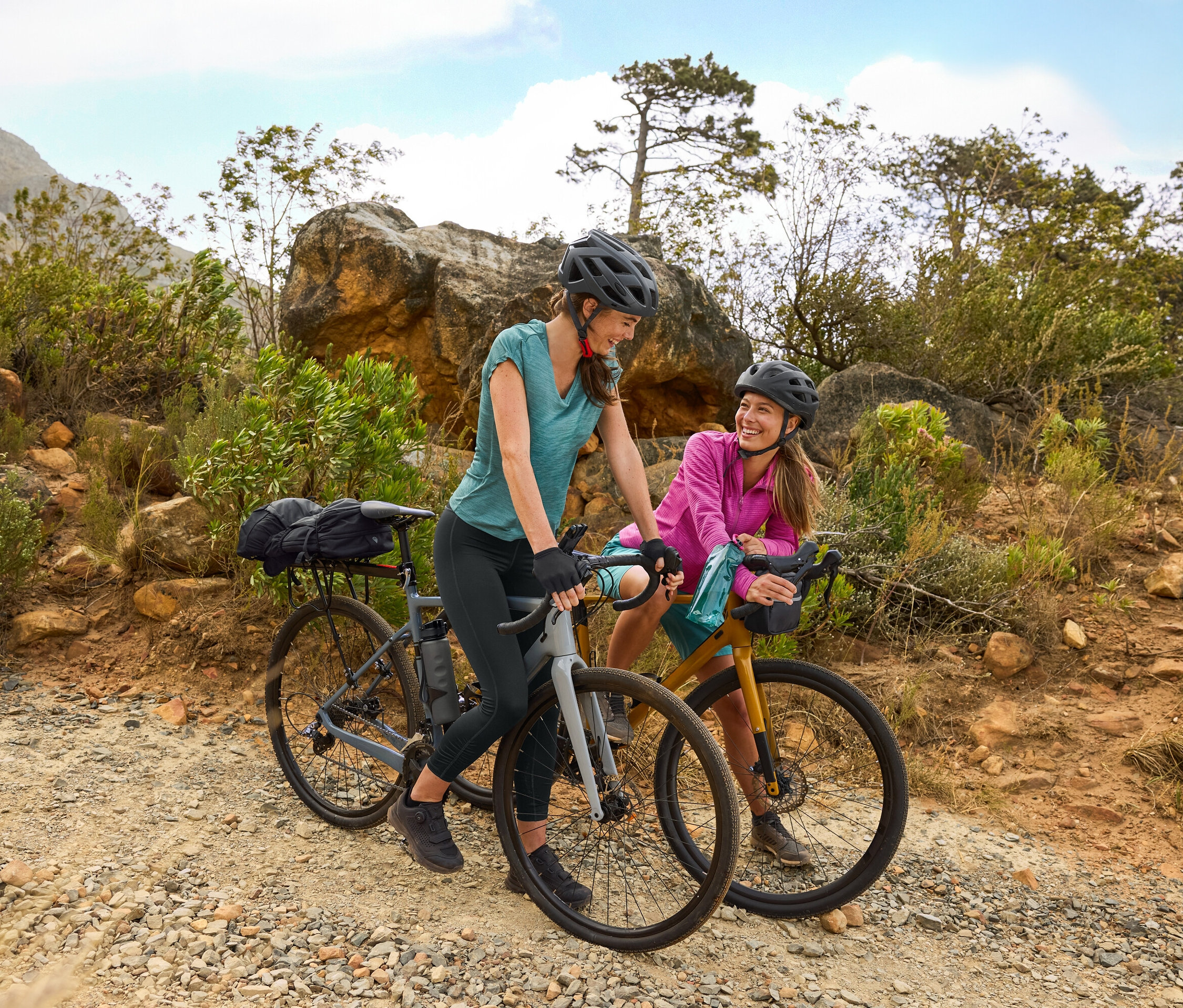 Dvě ženy s helmami stojí s koly na štěrkové cestě v kopcovitém terénu. Na kole je připevněna láhev na pití.